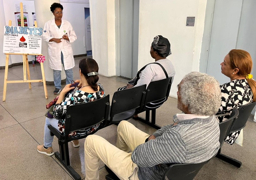 A foto mostra uma profissional de saúde falando a um grupo de pessoas sentadas na sala de espera de uma UBS; ela usa um jaleco branco e está posicionada ao lado de um cartaz onde se lê, em letras grandes, "Diabetes - sintomas" 